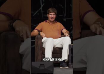 Entspannter Mann sitzt auf einem bequemen Sessel bei einer Veranstaltung, trägt ein orangefarbenes T-Shirt und weiße Hosen, zeigt eine lockere Haltung und lädt zum Austausch ein.