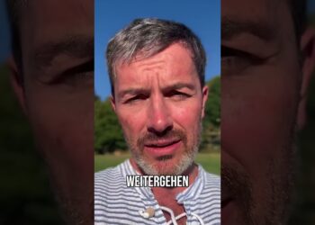 Man with short gray hair and beard, outdoors on sunny day, with the text "WEITERGEHEN" overlayed, in a casual striped shirt, demonstrating determination.